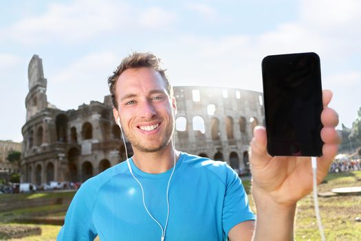 Smiling young runner showing smartphone with blank screen against Colosseum. Confident male jogger is listening music on sunny day. The empty space on mobile phone can be used for advertisement.