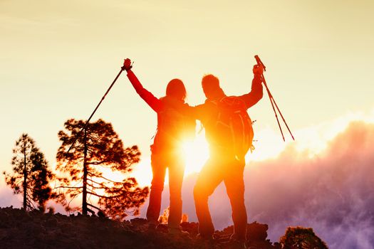 Successful hiking couple standing on mountain top during sunset cheering in success. Rear view of male and female hiker raising hiking poles against sky. Man and woman are celebrating during vacation.