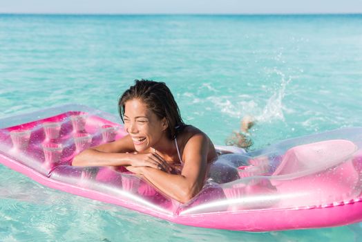 Beach girl having fun splashing water in ocean floating on pink pool float toy air mattress. Asian woman relaxing in the sun swimming in the perfect turquoise sea at holiday resort.