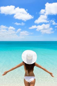 Happy beach vacation carefree woman on luxury tropical getaway destination wearing a white sun hat from behind with arms raised in freedom. Person from behind with slim body. Happiness and success.