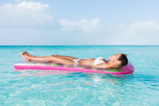 Beach sexy woman relaxing during suntan sunbathing on floating pink pool inflatable plastic air mattress float in pristine turquoise ocean water background at luxury destination getaway.
