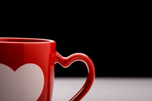 A burgundy coffee mug with heart shaped handle on white table with dark background.