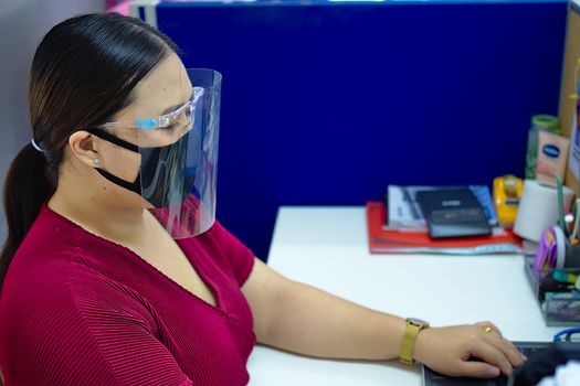 A young and beautiful Asian woman wearing a surgical mask and face shield to protect against Covid-19 while working in an office.