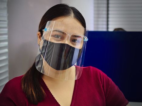 A young and beautiful Asian woman wearing a surgical mask and face shield to protect against Covid-19 while working in an office.