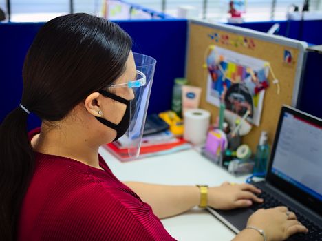 A young and beautiful Asian woman wearing a surgical mask and face shield to protect against Covid-19 while working in an office.