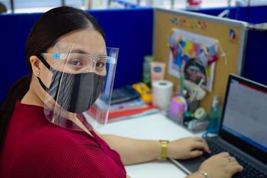 A young and beautiful Asian woman wearing a surgical mask and face shield to protect against Covid-19 while working in an office.