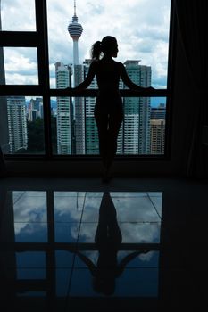 A slender girl is looking at the city through a large window. High floor