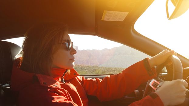 Woman driving a car. Young woman is driving a car on a sunny day. Confident woman in sunglasses driving car. Woman in a red jacket driving with Lens optical flare