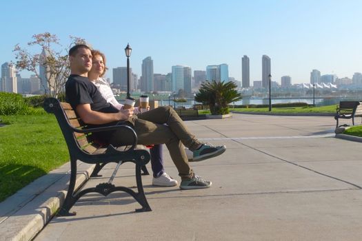Couple relaxing on San Diego City bench. In the background is the downtown San Diego with skyscrapers. Concept about love, relationship, and travel. Young couple resting on the promenade in the city.