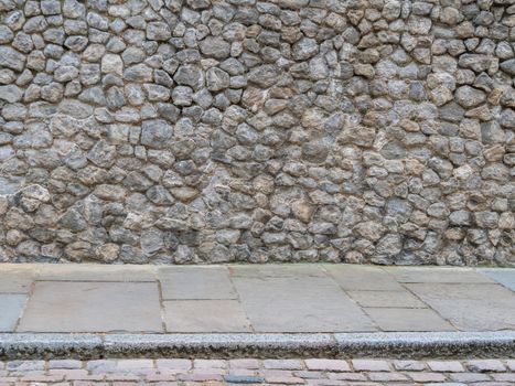 Natural rough stone wall texture background with concrete slabs and a curb on the floor. Stone wall background. Background of stone wall of boulders and pebbles. Cobble stone wall texture.