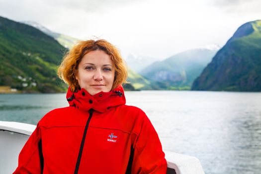 Young beautiful woman in a red jacket on the background of Norwegian fjord. Journey through the Norwegian fjords. Red-haired freckled young woman in red jacket. Tourist concept. Journey on the ship.