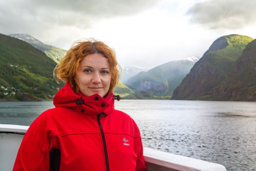 Young beautiful woman in a red jacket on the background of Norwegian fjord. Journey through the Norwegian fjords. Woman on the deck of a ship in Norway. Red-haired freckled young woman in red jacket.