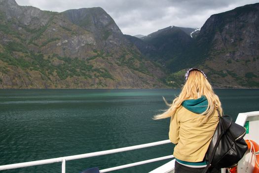 Young woman with a backpack on the deck of the ferry looks at the Norwegian fjords. Woman blonde tourist on a ship admires the nature of Norway. A young woman travels alone. Tourist concept.