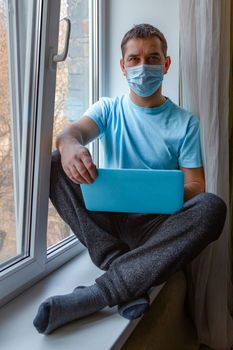 Young man in a blue protective medical mask sits on a windowsill with a laptop and looks at the camera. Freelance work, working at home, online learning, studying, lockdown concept. Distance education