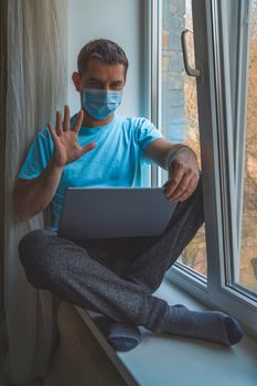 Man Greets and Talks at Video Chat using Laptop. Man in a protective medical mask sits on a windowsill with a laptop and talks online. Stay at home concep. Coronavirus covid-19 social distancing.