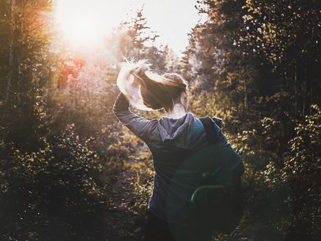 view of beautiful woman traveler standing in forest, Estonia. lifestyle tourist girl backpack straightening her hair in sunset time. Vacation concept, summer time. Toned image. copy space
