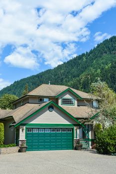 Big family house with wide concrete driveway, green wide garage door, decorated front yard. Residential house with mountain and blue sky background