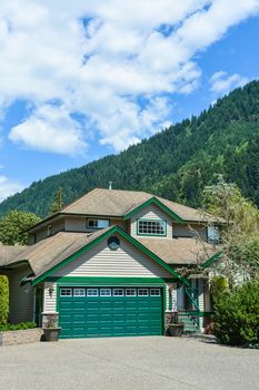 Big family house with wide concrete driveway, green wide garage door, decorated front yard. Residential house with mountain and blue sky background