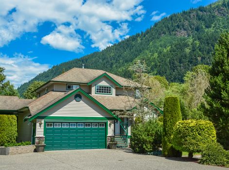 Big family house with wide concrete driveway, green wide garage door, decorated front yard. Residential house with mountain and blue sky background