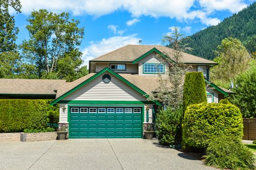 Big family house with wide concrete driveway, green wide garage door, decorated front yard. Residential house with mountain and blue sky background