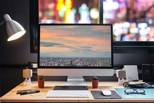 Graphic designer's workspace with city night view with a pen tablet, a computer and cityscape background for text