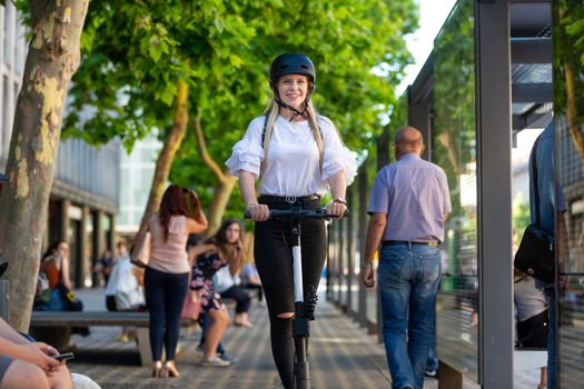 Trendy fashinable teenager, beautiful blonde girl riding public rental electric scooter in urban city environment. Eco-friendly modern public city transport.