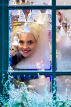 A young blonde woman looks through a window pane that is frozen and adorned with Christmas decorations. The concept of Christmas time - image
