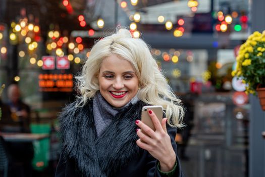 Young, beautiful blonde woman taking selfies against the background of festive lights. Christmas time concept. Copy space - image
