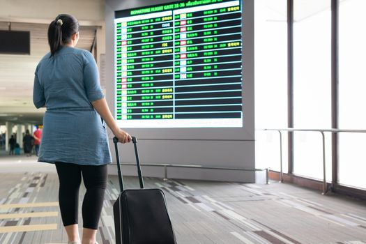 Woman with luggage wait and look at terminal gate information board checking for her flight in international airport. Suitcases in the departure lounge, holiday vacation concept, traveler suitcases