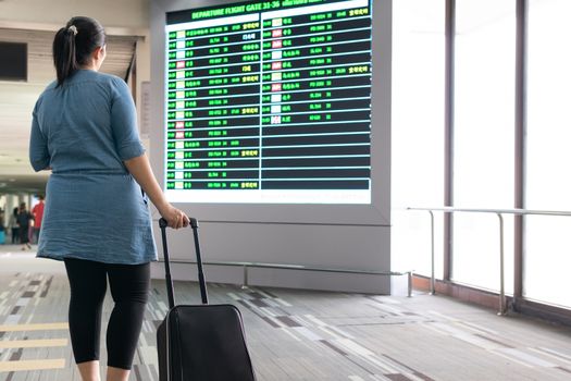 Woman with luggage wait and look at terminal gate information board checking for her flight in international airport. Suitcases in the departure lounge, holiday vacation concept, traveler suitcases