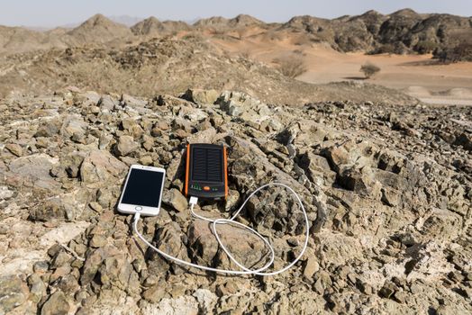 Smartphone charging with solar power bank with Dual USB port. These gadgets are lying on rocks, in the deserted mountain with some sand dunes in background.
