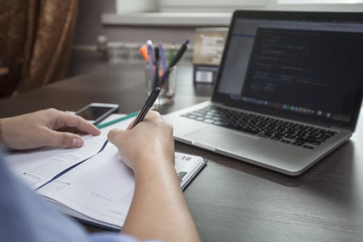 Man's hands make entries in the notebook, an open laptop in the face of