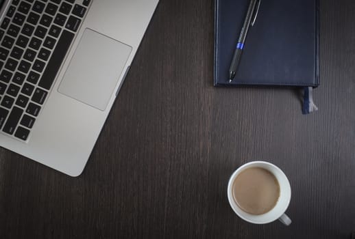 On a wooden table a laptop, a cup of coffee and a notebook