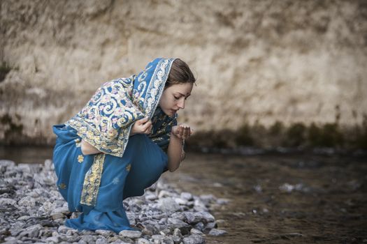 The girl in the Indian sari sits at the small river and drinks from hands