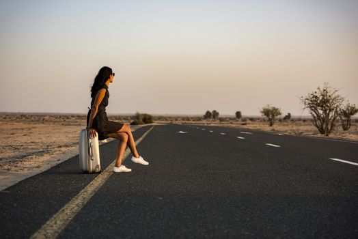 Woman waiting with her luggage on rural road in the desert for a lift or a vehicle to come