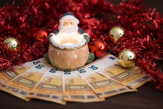 Christmas money business concept: a statuette of Santa Claus with lit little candle on some fifty euro banknotes with red and gold baubles and wreath decoration with bokeh effect on light wooden table