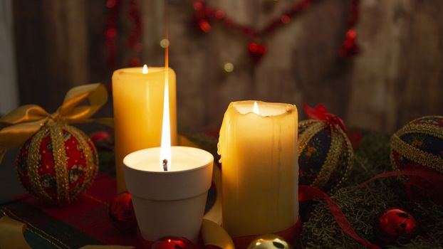 Close up of a Lit candle with big flame on Christmas table cloth with around pine branches, decoupage baubles, with lit candles and hanging Christmas decoration on wooden background with bokeh effect