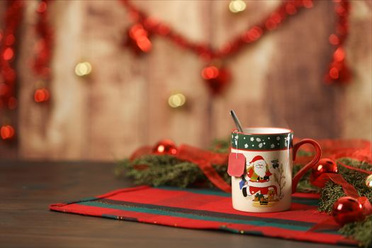 Christmas mug with hanging empty tea label on Christmas table cloth with around pine branches, red baubles and hanging Christmas decoration on wooden background with bokeh effect