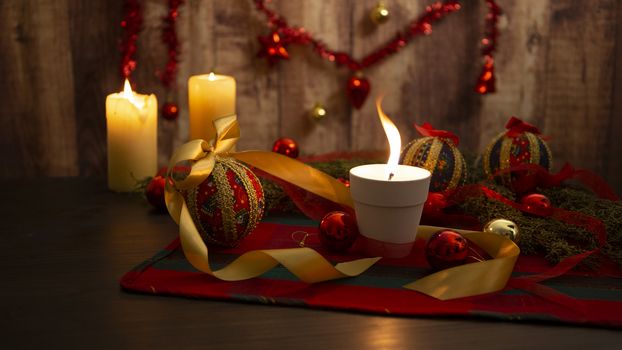 Lit candle with big flame on Christmas table cloth with around pine branches, decoupage baubles, with lit candles and hanging Christmas decoration on wooden background with bokeh effect