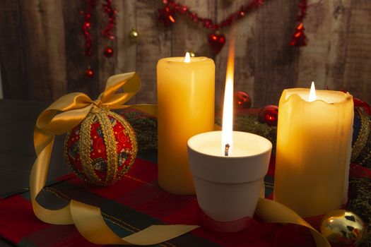 Close up of a Lit candle with big flame on Christmas table cloth with around pine branches, decoupage baubles, with lit candles and hanging Christmas decoration on wooden background with bokeh effect