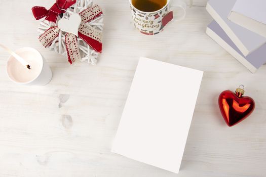 Christmas view from top for a book presentation: book with blank cover with a white lit candles, Christmas decorated cup of tea, red heart bauble, a shabby decorated white heart on light wooden table