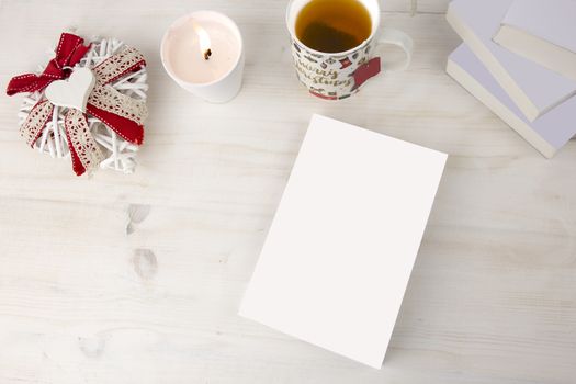 Christmas view from top for a book presentation: book with blank cover with a white lit candles, Christmas decorated cup of tea other three books, a shabby decorated white heart on light wooden table