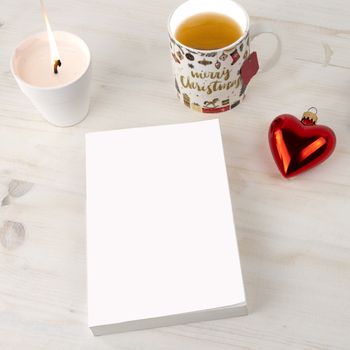 Christmas setting view from top for a book presentation: book with blank cover with a white lit candles, Christmas cup of tea and red baubles with heart shape on light wooden table