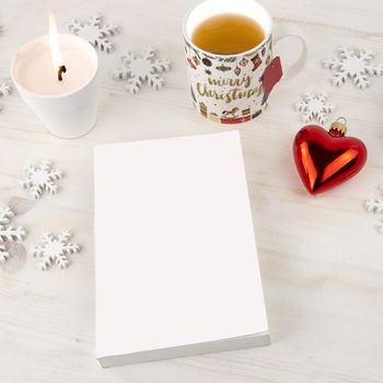 Christmas setting view from top for a book presentation: book with blank cover with a white lit candles, wooden snowflakes, Christmas cup of tea and red baubles with heart shape on light wooden table