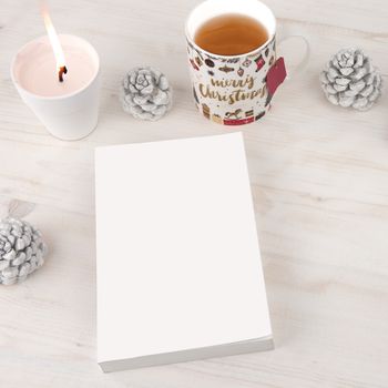 Christmas setting view from top for a book presentation: book with blank cover with a white lit candles, Christmas decorated cup of tea and white pine cones on light wooden table