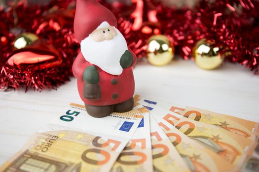 Christmas money business concept: a statuette of Santa Claus on some fifty euro banknotes with red and gold baubles and wreath decoration with bokeh effect on light wooden table