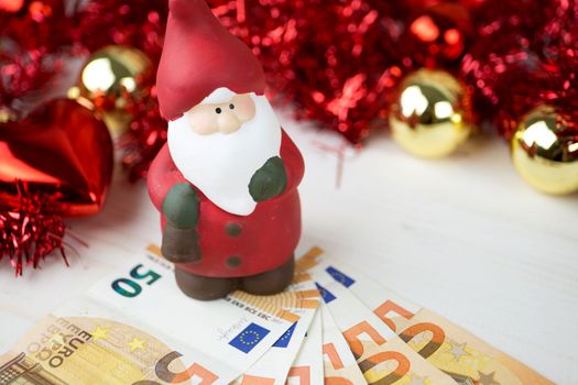 Christmas money business concept: a statuette of Santa Claus on some fifty euro banknotes with red and gold baubles and wreath decoration with bokeh effect on light wooden table