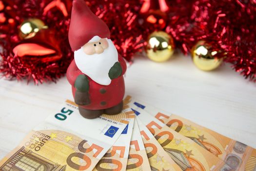 Christmas money business concept: a statuette of Santa Claus on some fifty euro banknotes with red and gold baubles and wreath decoration with bokeh effect on light wooden table