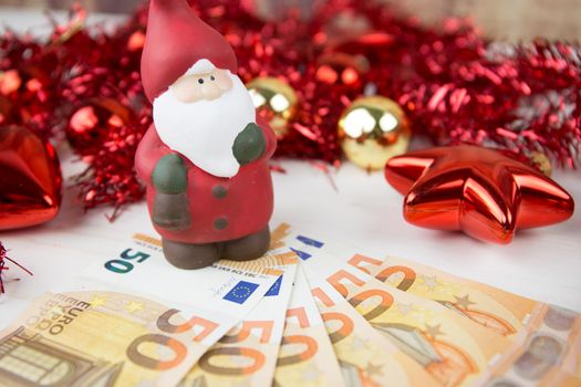 Christmas money business concept: a statuette of Santa Claus on some fifty euro banknotes with red and gold baubles and wreath decoration with bokeh effect on light wooden table