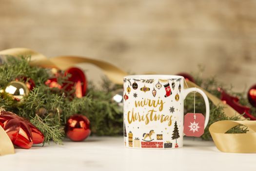 Close up of a Christmas mug with hanging empty tea label with around pine branches, red baubles and satin gold ribbon on wooden background with bokeh effect